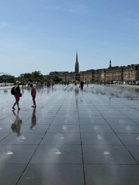 People walking across the miroir d&rsquo;eau in Bordeaux on a sunny day, with mist and the Saint-Michel spire in the background